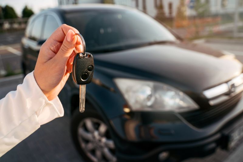 Womans hand holding a car key with a black car in the background - Do you Need to Hire a Car in Barcelona?